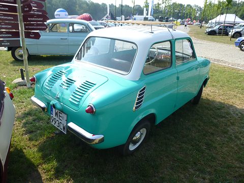 Turquoise small car with white roof from the rear three-quarter on grass, tents and cars beyond. Opens a larger view