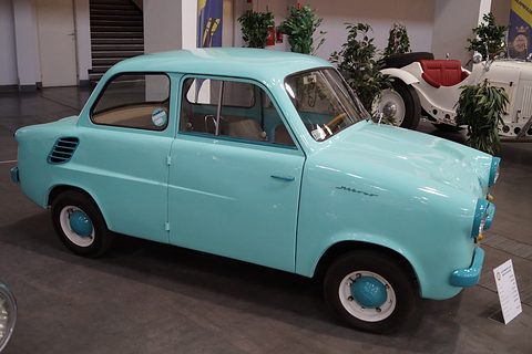 Turquoise small two-door saloon on a dark floor among potted plants and a white vintage roadster. Opens a larger view