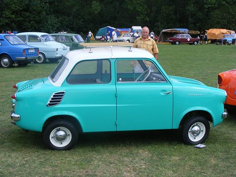 Turquoise small saloon with a white roof on grass, tents and classic cars behind. Opens a larger view
