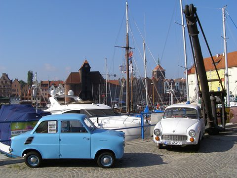 A small blue car and a white saloon on cobbles among moored yachts and historic waterfront buildings. Opens a larger view