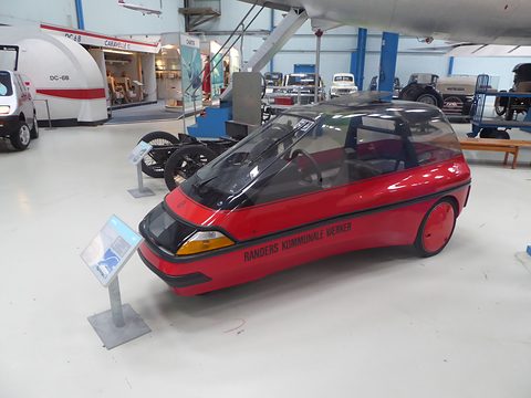 Red and black wedge-shaped single-seater with a smoked canopy, beside an information stand in a large museum hall. Opens a larger view