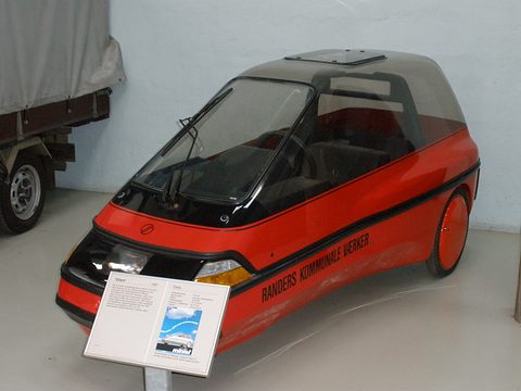 A red and black single-seat electric three-wheeler with a tinted canopy, an information board standing before it. Opens a larger view