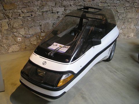 White and black single-seat three-wheeler with a smoked dome canopy against a rough stone wall, a leaflet on its dashboard. Opens a larger view