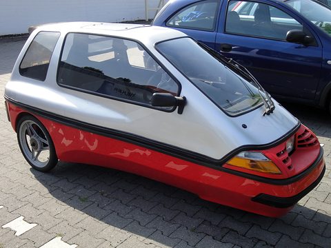 Silver three-wheeler with red lower panels and flame graphics, its long glazed cabin beside a dark blue hatchback. Opens a larger view