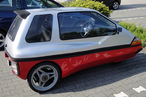 Rear three-quarter of a silver and red three-wheeler with a roof spoiler, alloy wheel and steering wheel visible inside. Opens a larger view