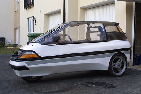 Side view of a white and grey wedge-shaped three-wheeler with a long glazed cabin and alloy wheel, outside garages. Opens a larger view