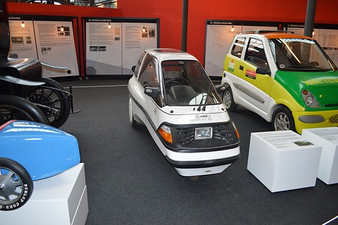 White single-seat three-wheeler with a black grille marked ‘mini-el City’ beside a green and yellow microcar on a dark floor. Opens a larger view