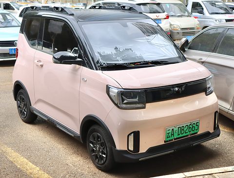 Pale pink boxy city car with a black roof, dark wheels and slim headlamps, parked among other cars. Opens a larger view