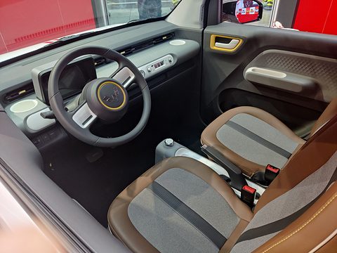 Grey and brown seats, a two-spoke steering wheel and a grey dashboard with a yellow door handle, seen from above. Opens a larger view