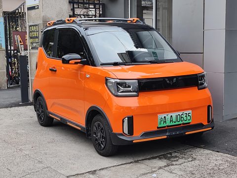 Orange boxy city car with a black roof and roof rails, parked on a pavement by a doorway. Opens a larger view
