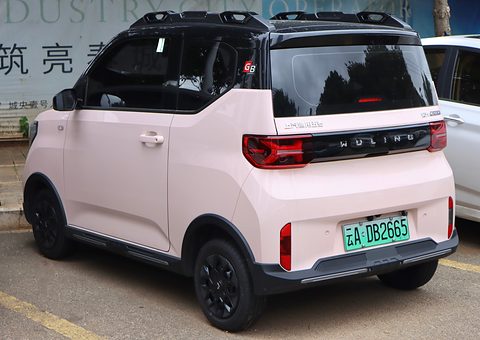 Rear of a pale pink boxy city car with a black roof, roof rails and dark wheels, parked on tarmac. Opens a larger view