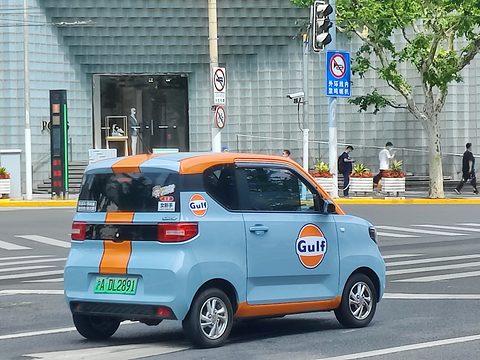 Rear of a pale blue boxy city car with an orange stripe and Gulf roundels, crossing a road junction.