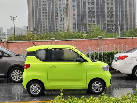 Side view of a bright lime-green boxy three-door city car on a wet road, tower blocks behind. Opens a larger view