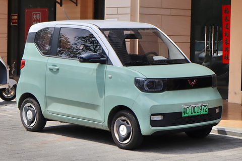 Pastel-green boxy city car with a white roof and round-cornered lamps, parked on a pavement by a shopfront.
