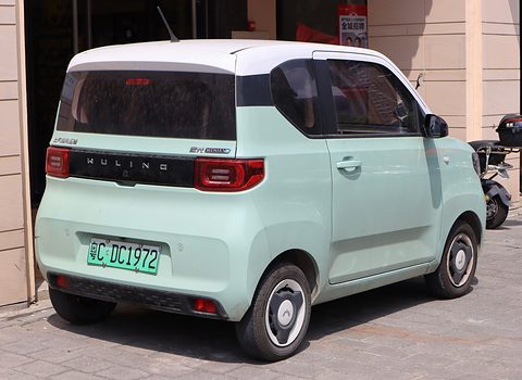 Rear of a pastel-green boxy city car with a white roof and Wuling lettering on a black band, parked by a shop. Opens a larger view