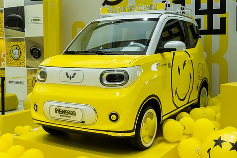 Bright yellow boxy city car with a smiley-face graphic, among yellow balloons on a yellow promotional stand. Opens a larger view
