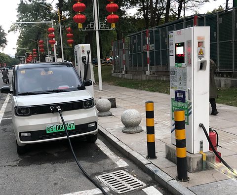 White boxy city car connected by a cable to a white charging post on a pavement hung with red lanterns.