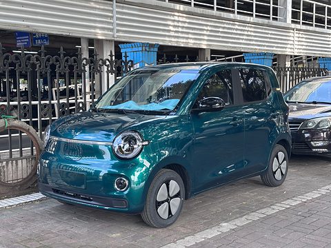Dark green rounded small hatchback with chrome-ringed round lamps parked on paving by a railing. Opens a larger view