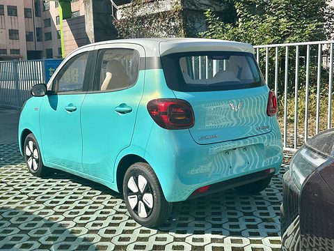 Rear of a turquoise rounded small hatchback with red tail lamps and a white roof, on grass pavers. Opens a larger view