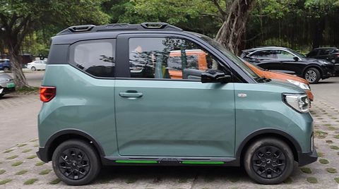 Side view of a grey-green boxy city car with a black roof and rails and black wheels, in a car park. Opens a larger view