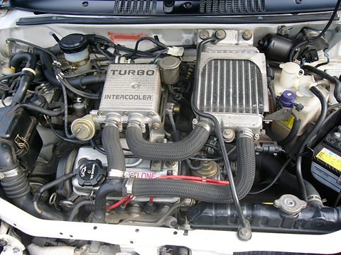 Engine bay with a silver TURBO INTERCOOLER cover, hoses and a white radiator support. Opens a larger view