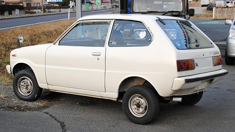 Rear three-quarter of a white small hatchback on a cracked tarmac lot. Opens a larger view
