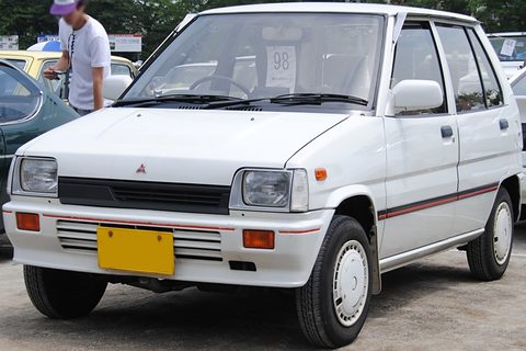 White small hatchback with a yellow plate seen from the front, a man standing behind. Opens a larger view