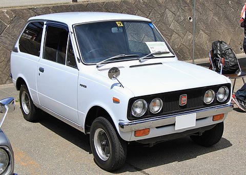White small hatchback-shaped saloon with four round headlamps, parked by a concrete wall. Opens a larger view