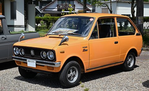 Orange small car with quad headlamps parked on gravel, houses behind.