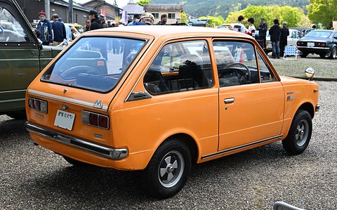 Rear three-quarter of an orange small car on gravel, visitors in the distance. Opens a larger view