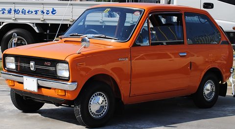 Orange small car with round headlamps and a white plate, a lettered truck behind. Opens a larger view