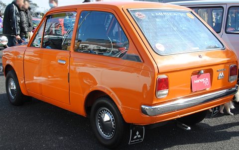 Rear three-quarter of an orange small car with a MINICA plate, visitors in coats behind. Opens a larger view