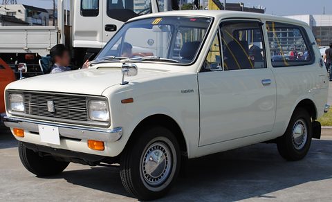 White small estate van with a grille between round headlamps, a truck and people behind. Opens a larger view