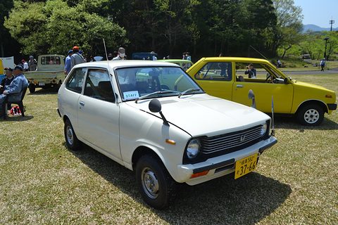 White small hatchback on grass, a yellow hatchback and visitors behind. Opens a larger view