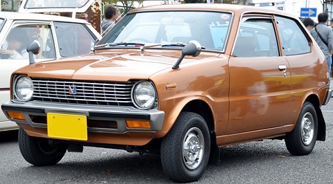 Copper-brown small hatchback with round headlamps and a yellow plate, visitors behind. Opens a larger view