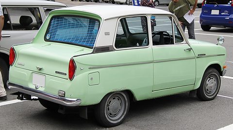 Rear three-quarter of a pale green saloon with a white roof and a slatted sun blind on its rear window. Opens a larger view