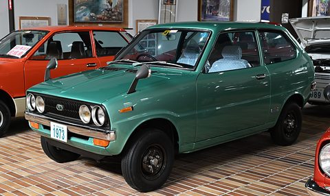 Teal-green small hatchback on a herringbone brick floor, an orange car behind. Opens a larger view