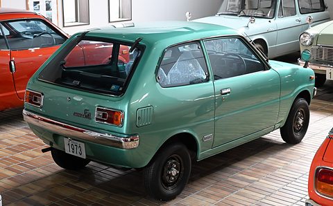 Rear three-quarter of a teal-green small hatchback on a herringbone floor. Opens a larger view
