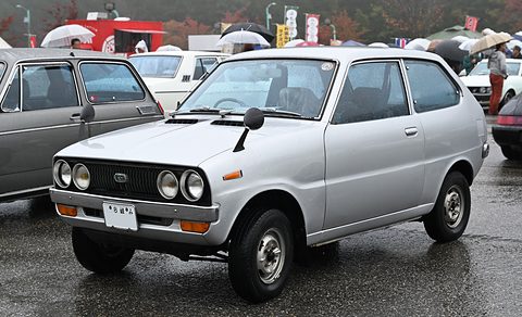 Silver small hatchback with round headlamps on wet tarmac, umbrellas behind. Opens a larger view