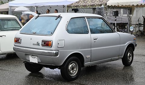 Rear three-quarter of a silver small hatchback under a striped awning, raindrops on its roof. Opens a larger view