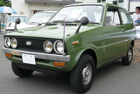 Olive-green small hatchback with round headlamps, other cars and a visitor behind. Opens a larger view