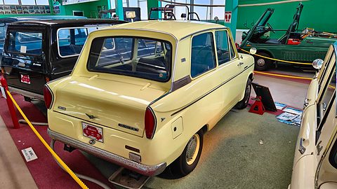 Rear three-quarter of a cream small saloon with fins and round tail lamps, green walls behind. Opens a larger view