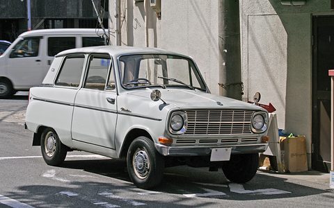 White small saloon with a wide slatted grille and round headlamps, parked by a grey building. Opens a larger view