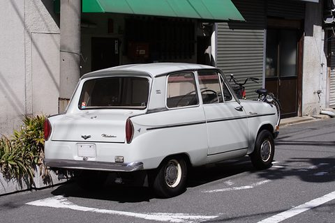 Rear three-quarter of a white small saloon with a near-vertical rear window, plants and a shop front behind.