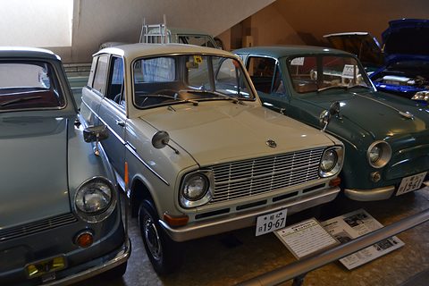 Pale grey small saloon between a blue-grey car and a dark green car in a museum. Opens a larger view