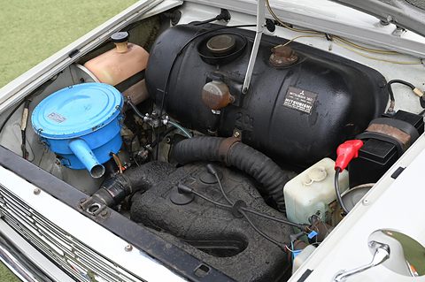 Open engine bay with a blue air cleaner, a black fuel tank and hoses on a white car. Opens a larger view