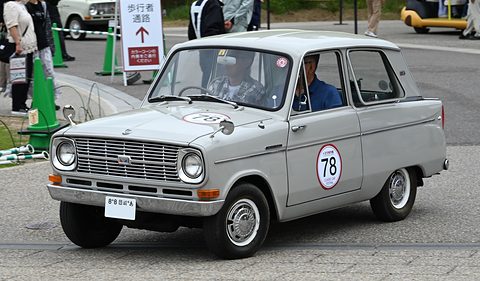Pale grey small saloon with a number 78 roundel on the door and two occupants, a sign behind. Opens a larger view
