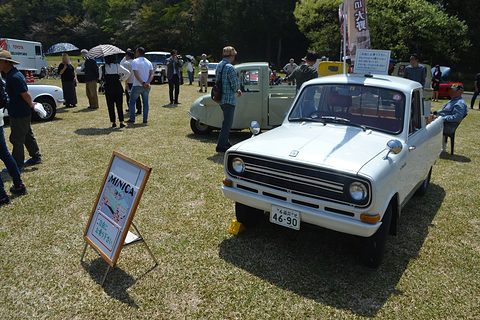 White small pickup on grass beside a wooden board, visitors and umbrellas behind. Opens a larger view