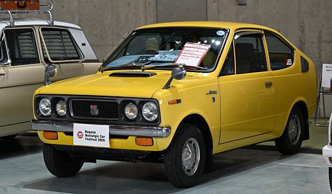 Yellow small coupé with four headlamps and a show plaque on its bumper. Opens a larger view