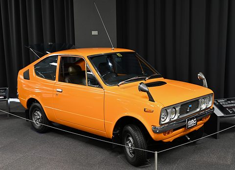Orange small coupé with twin headlamps on a dark floor in front of black drapes. Opens a larger view